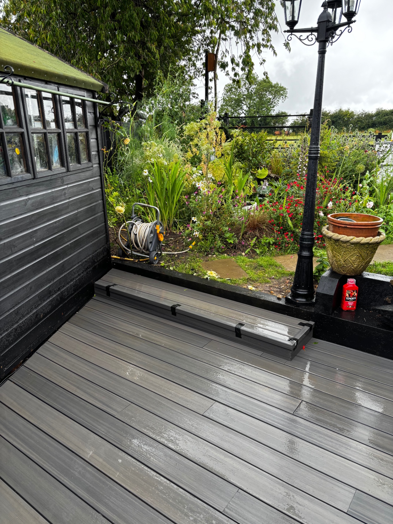Wide composite decking area with black shed, border planting and garden lighting