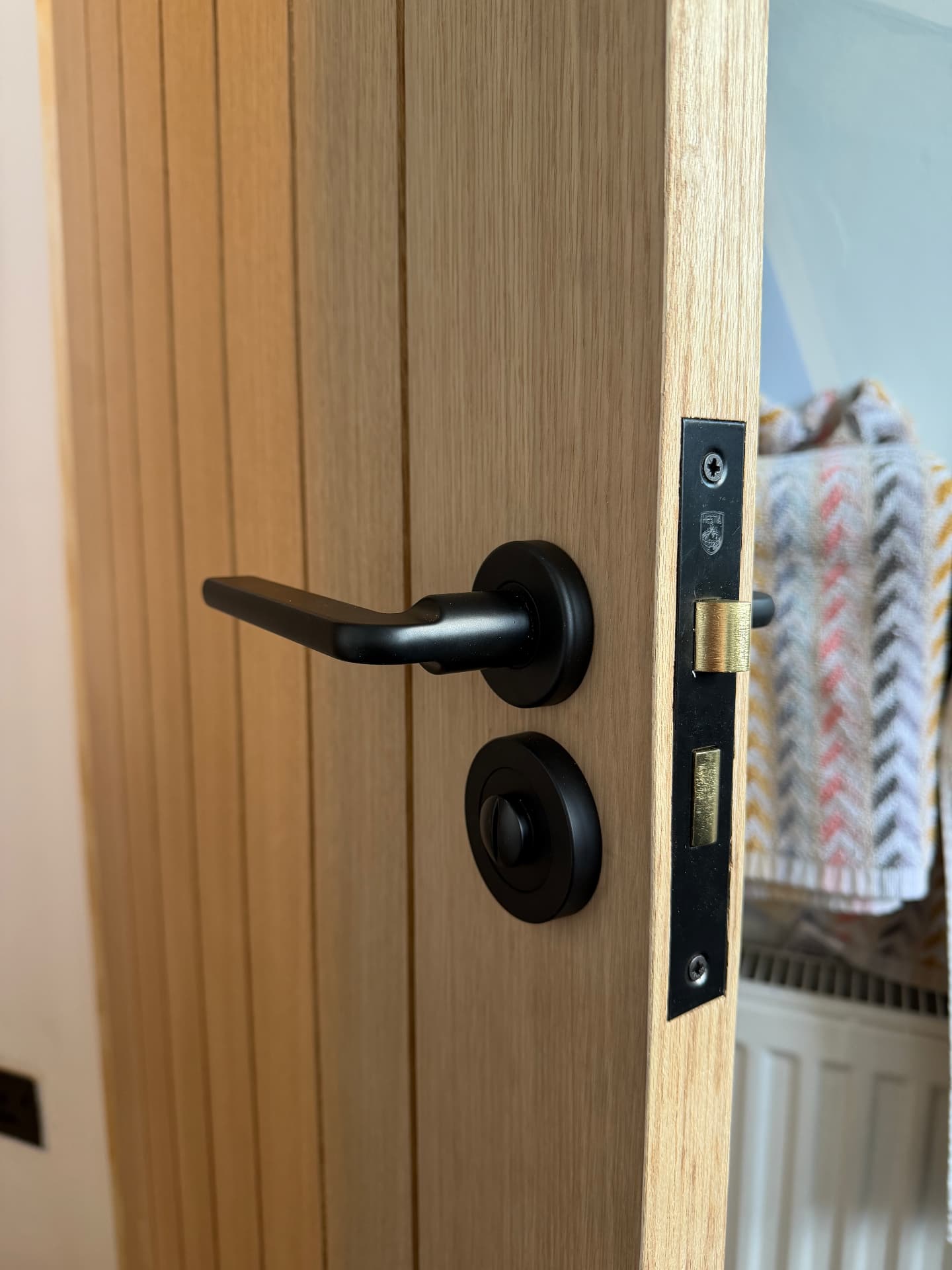 Matte black lever and thumbturn on a light oak slatted door, with the latch plate fitted in the door edge